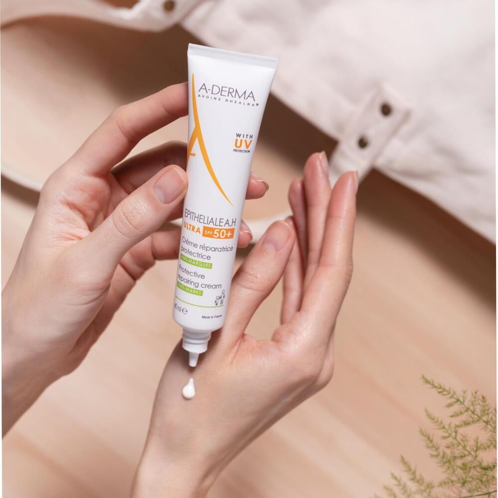 Mains tenant un tube A-DERMA EPITHELIALE A.H. ULTRA SPF 50+. Crème sort. Fond en bois.