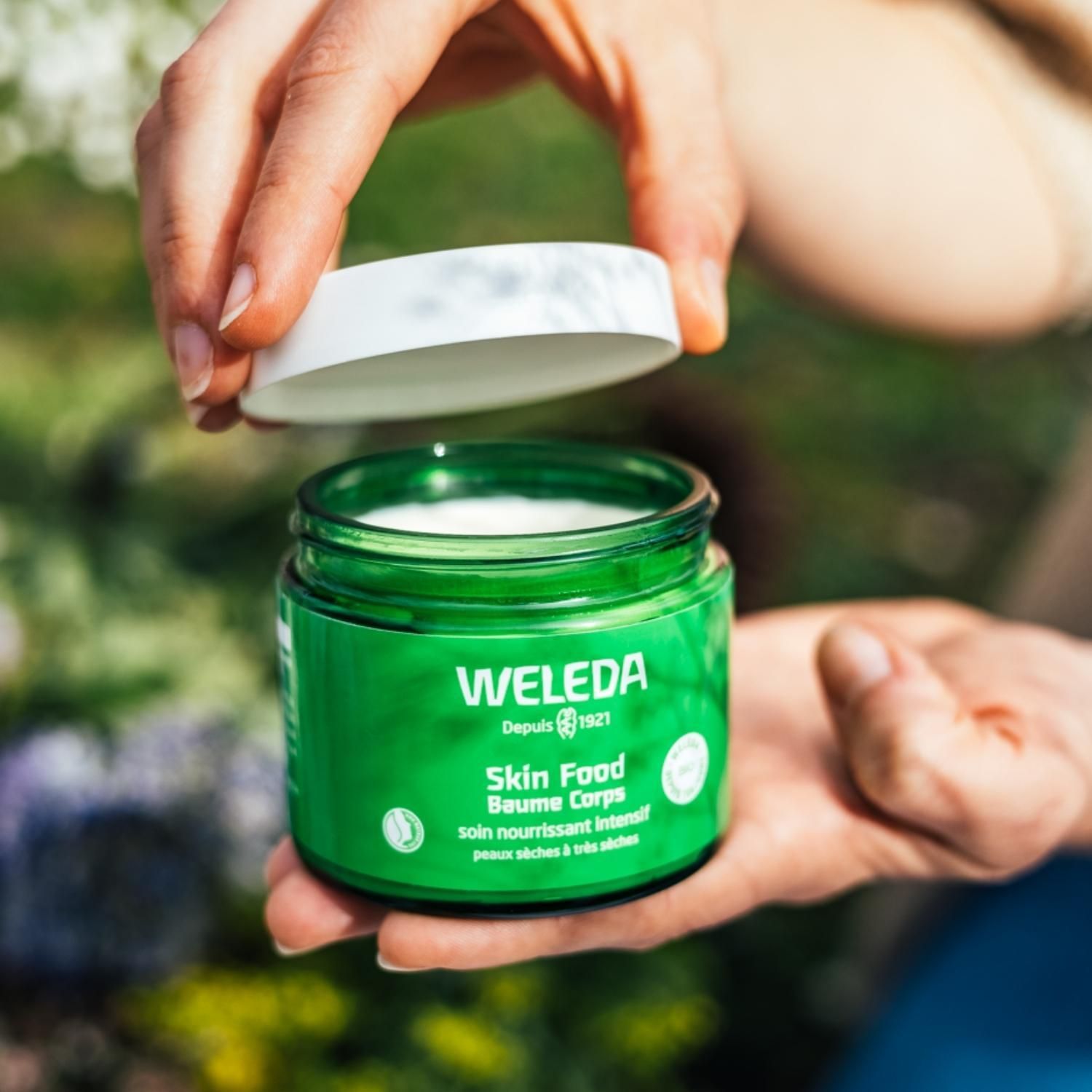 Hände halten einen geöffneten grünen Tiegel mit weißem Deckel. Sichtbar: Skin Food Baume Corps.