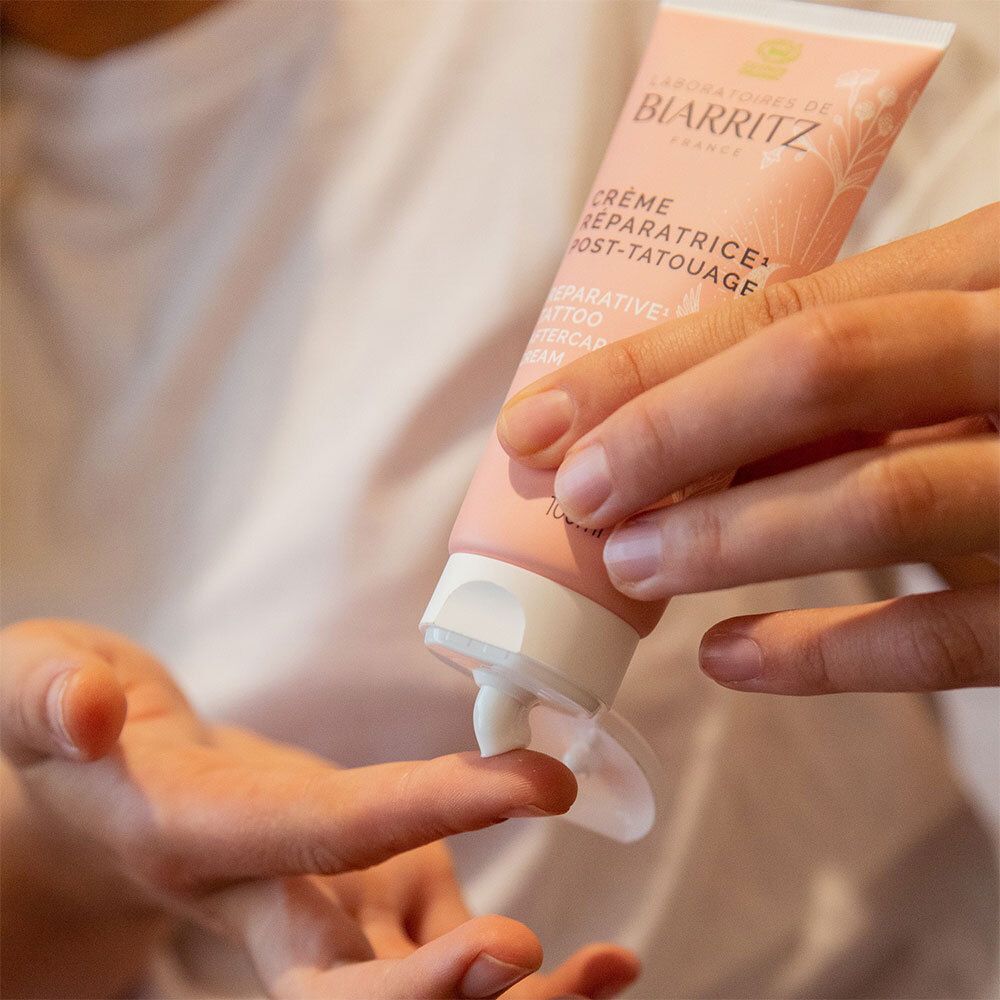Personne ouvrant le tube et appliquant la crème sur le doigt. Inscription: Crème Réparatrice Post-Tatouage. Marque: Laboratoires de Biarritz.