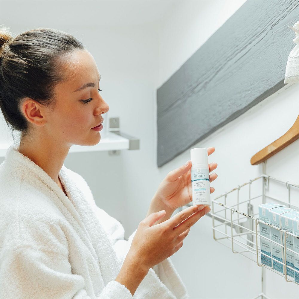 Femme en peignoir tenant un flacon. Étagère avec produits en arrière-plan.