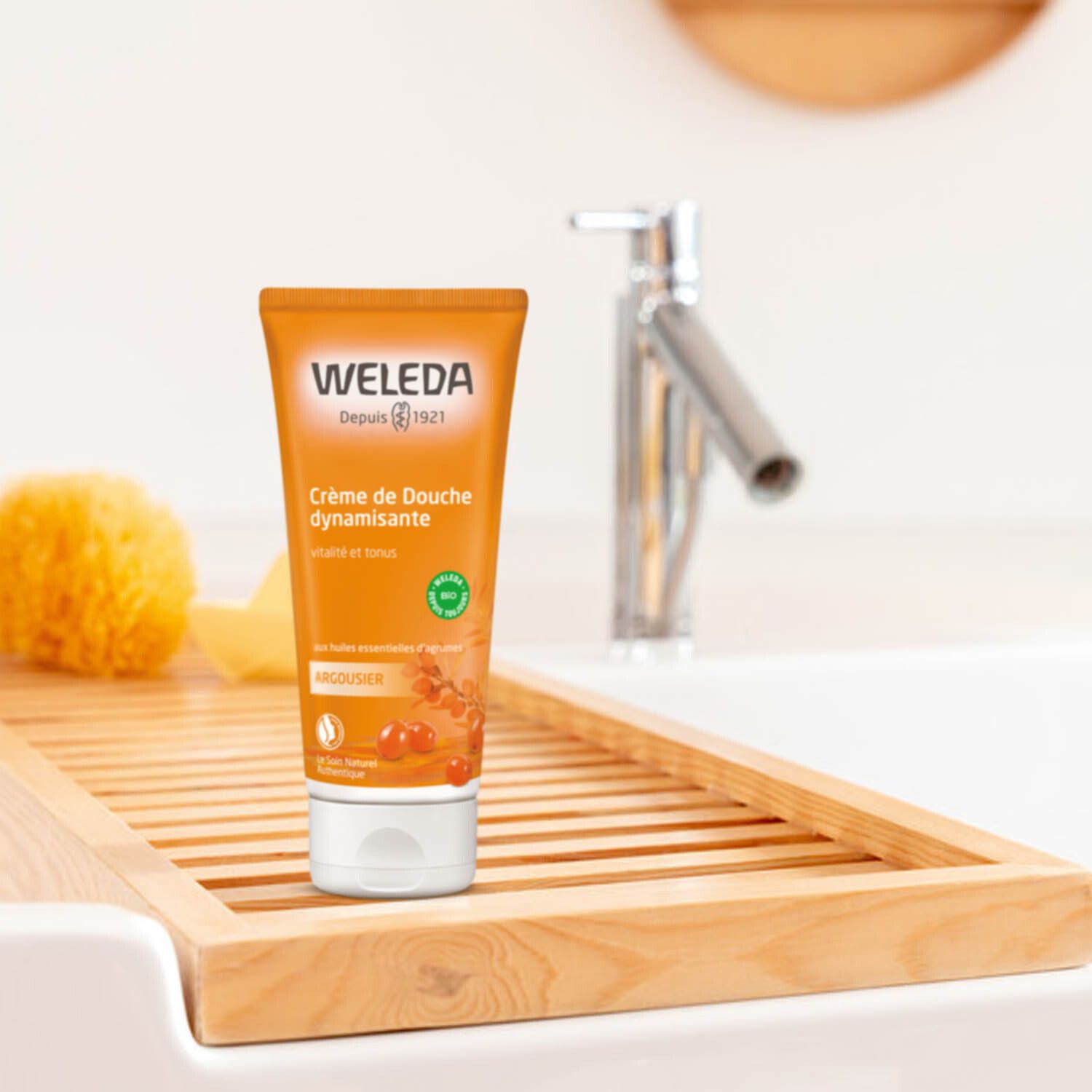 Tube orange près d'un lavabo. Marque: Weleda. Texte: Crème de Douche dynamisante. Label Bio. Sur une étagère en bois.