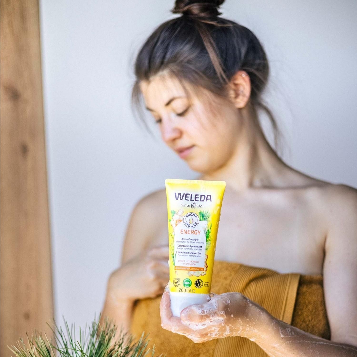 Femme tenant un tube jaune "WELEDA ENERGY Aroma-Duschgel". Elle est enveloppée dans une serviette.