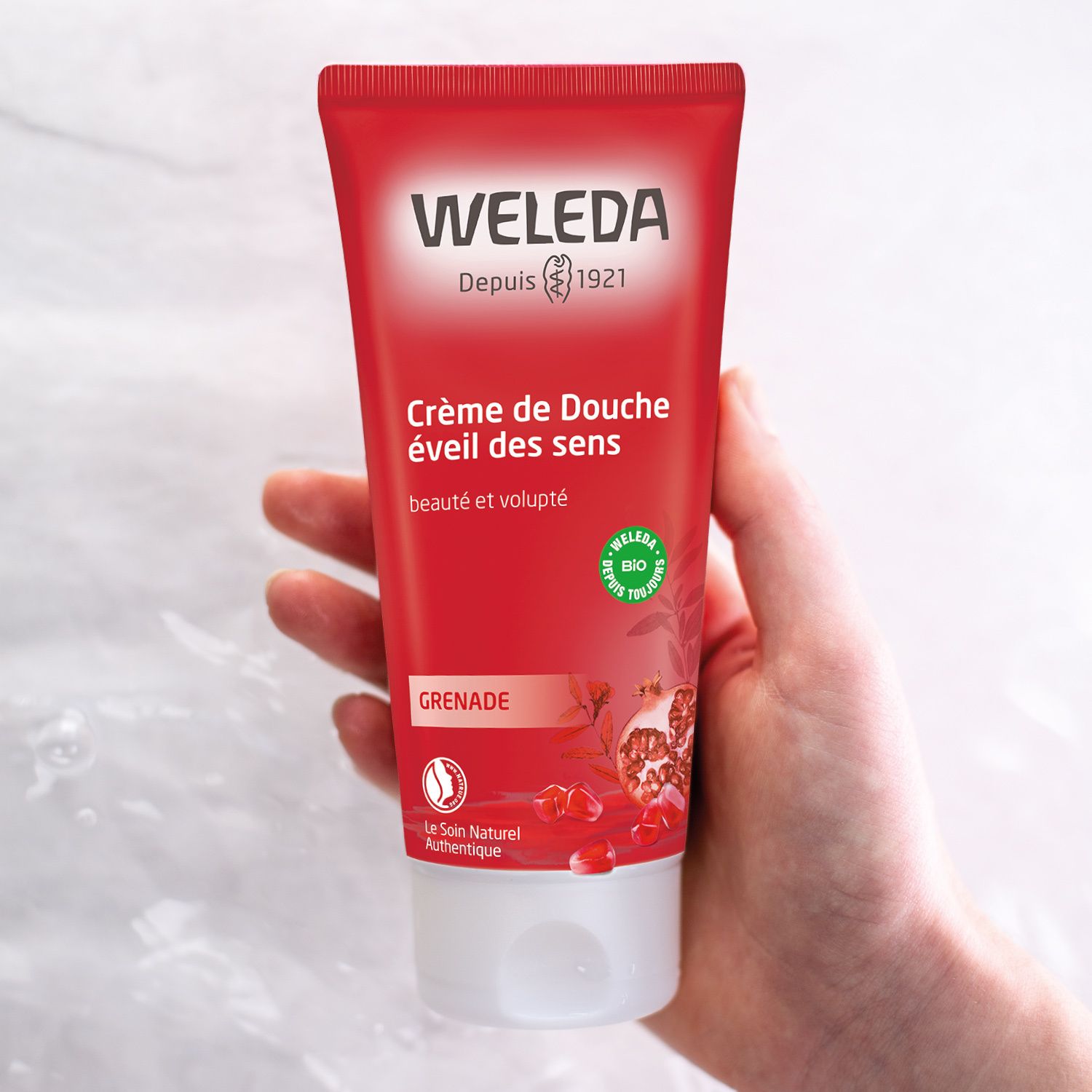 Tube rouge dans la main. Texte: Crème de Douche éveil des sens. Grenade. Logo. Fond blanc.