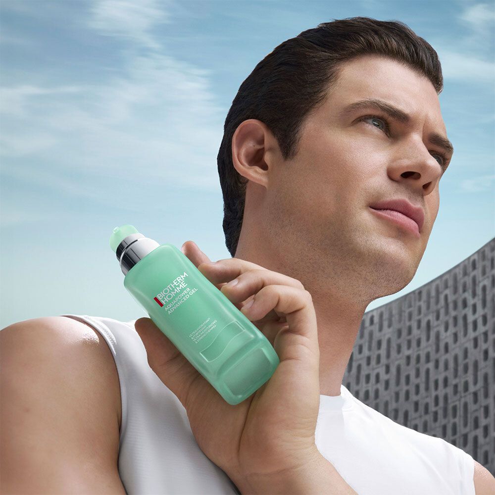 Mann hält BIOTHERM Homme Aquapower Gel. Türkisgrüne Flasche. Hintergrund: Himmel und Gebäude.