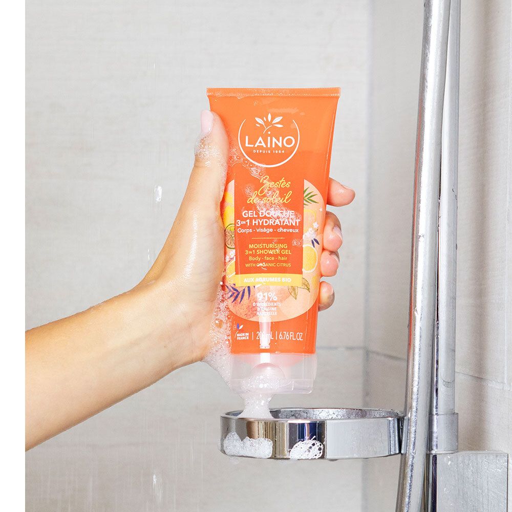 Hand hält orangefarbene Tube unter einer Duschbrause. Produktname und Logo sichtbar. Schaum um die Tube.