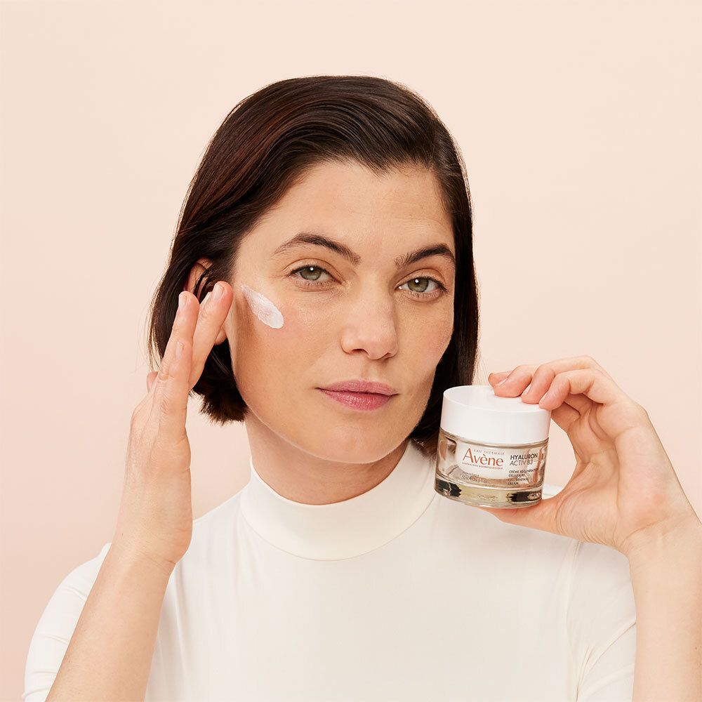 Femme appliquant de la crème. Elle tient un pot de crème. Nom du produit non visible.
