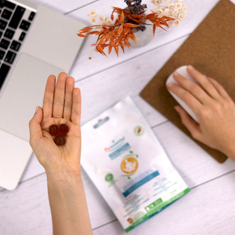 Personne tenant une main avec des gummies. À côté, un emballage du produit. Sur un bureau avec une souris et un ordinateur portable.