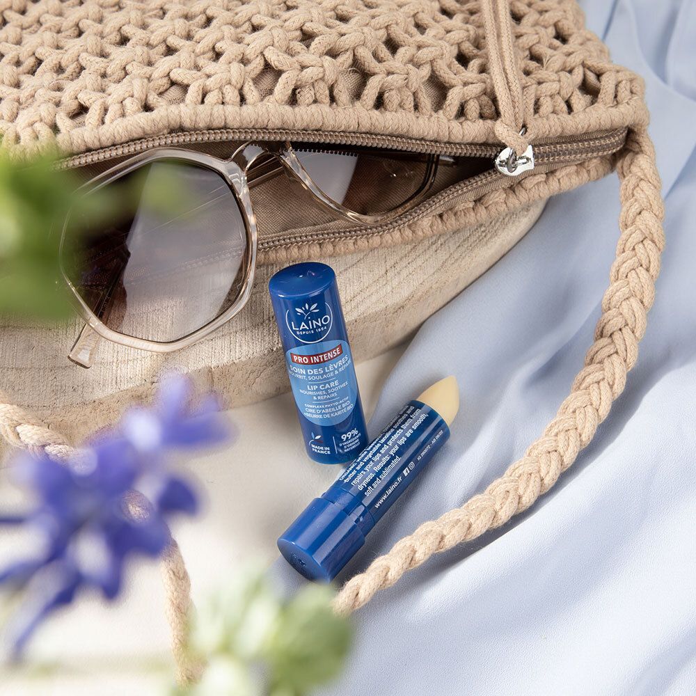 Zwei blaue Lippenpflegestifte mit LAINO-Logo in einer beigen Handtasche. Sonnenbrille daneben. Blaue Blume im Vordergrund.