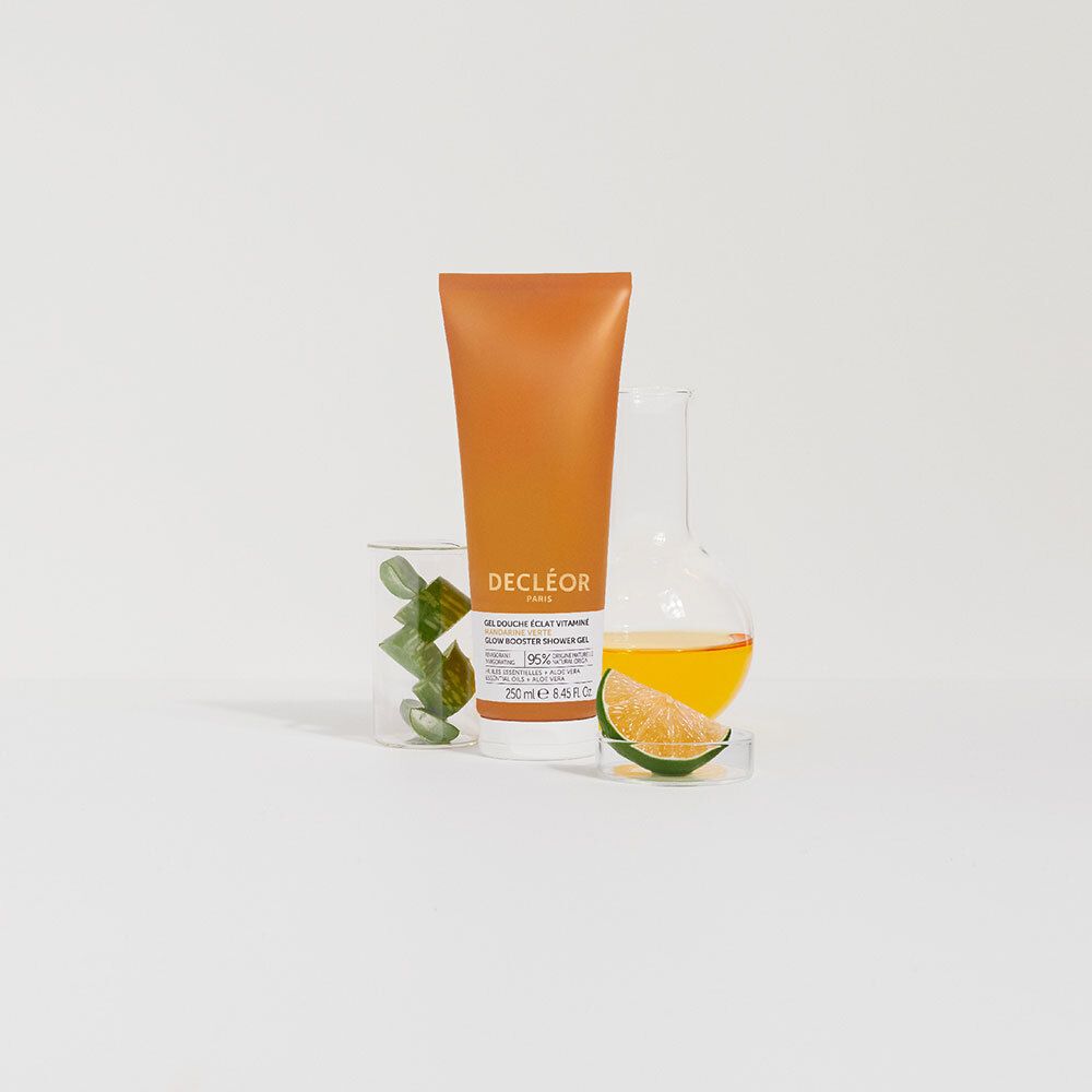 Oranje tube naast glazen containers met groene bladeren en vloeistof. Opschrift: DECLÉOR PARIS, GEL DOUCHE ÉCLAT VITAMINÉ.