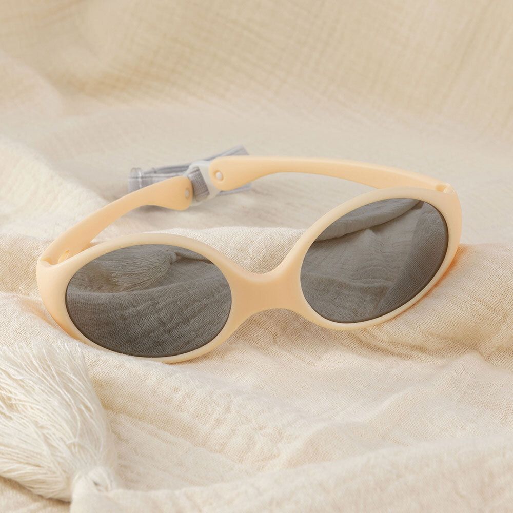Lunettes de soleil beiges avec verres gris et bandeau gris. Posées sur un tissu beige.