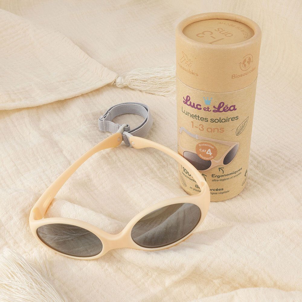 Lunettes de soleil beiges avec verres gris et bandeau gris. À côté, l'emballage. Inscription: Luc & Léa, lunettes de soleil 1-3 ans.