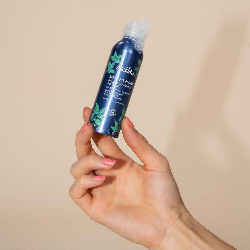 Hand hält eine Melvita Peelingpuder-Flasche. Blaue Flasche mit grünen Blättern.