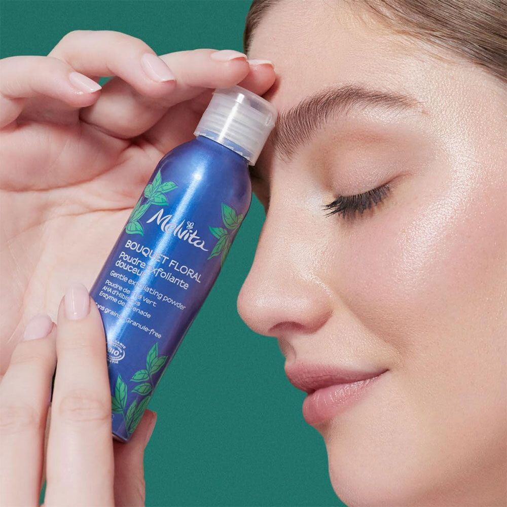 Melvita Peelingpuder-Flasche in weißem Pulvernebel. Blaue Flasche mit grünen Blättern.
