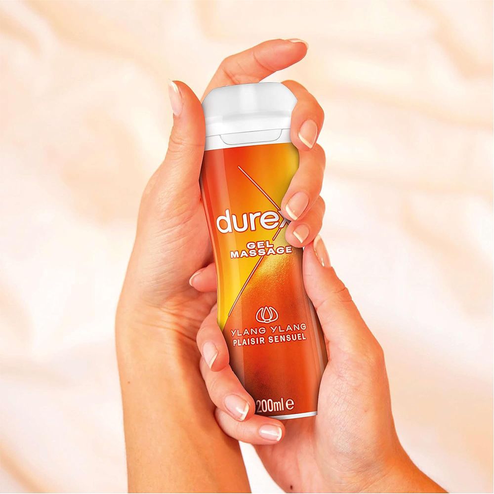 Mains tenant un flacon de gel de massage Durex. Étiquette orange-jaune. Texte : Gel Massage, Ylang Ylang, 200ml.
