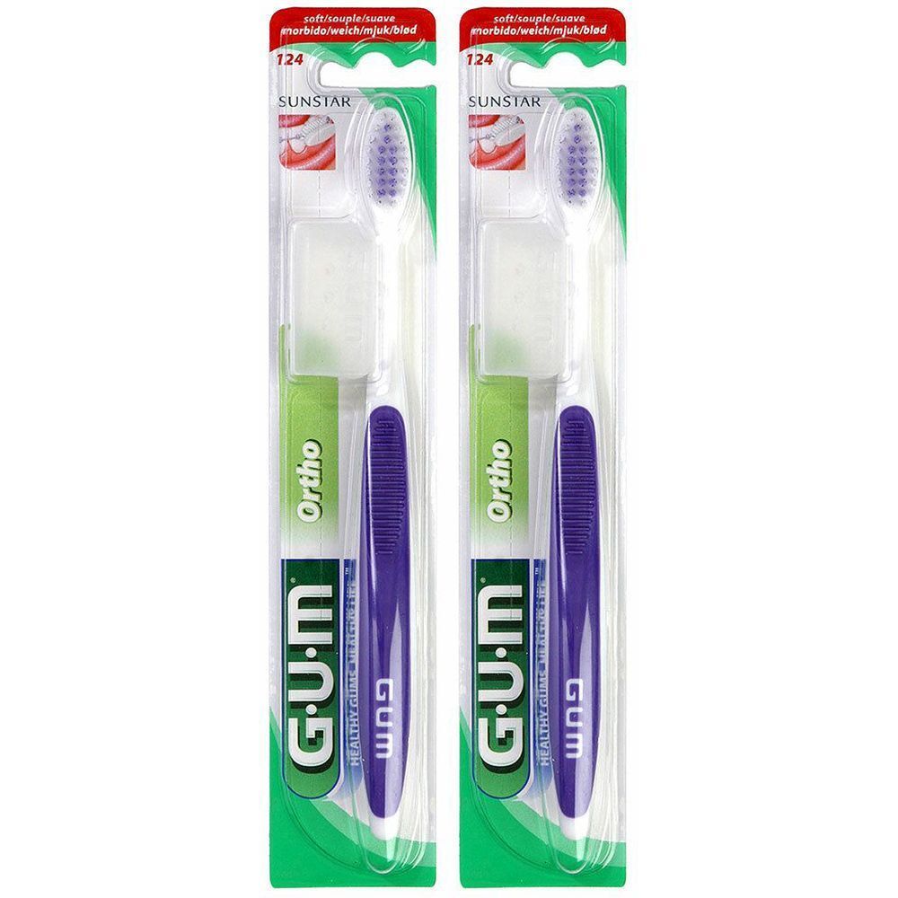 Deux brosses à dents emballées. Manche violet, tête blanche avec poils violets. Marque G.U.M, modèle Ortho. Emballage avec logo SUNSTAR.