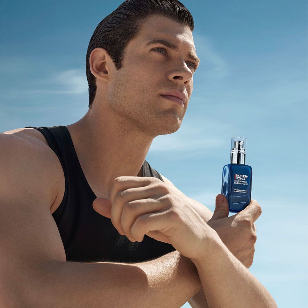 Homme tenant un flacon de sérum bleu. Fond : ciel. Il porte un débardeur noir.