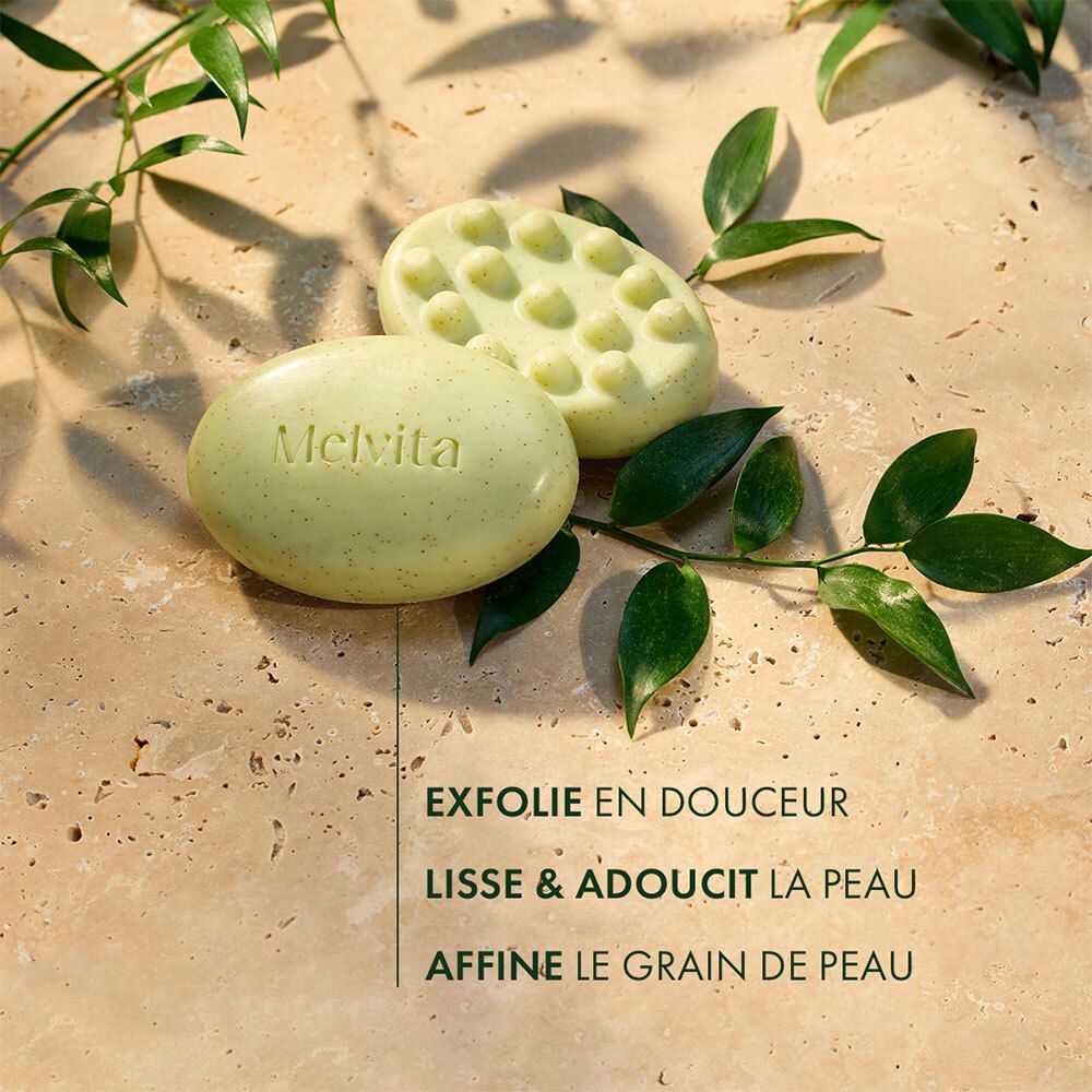 Zwei ovale Seifenstücke. Auf einem steht 'Melvita'. Text: Exfolie en douceur, Lisse & adoucit la peau, Affine le grain de peau.