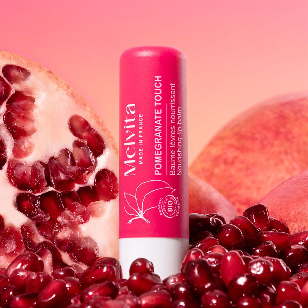 Melvita Lippenbalsam Pomegranate Touch umgeben von Granatapfelkernen und Fruchtstücken.