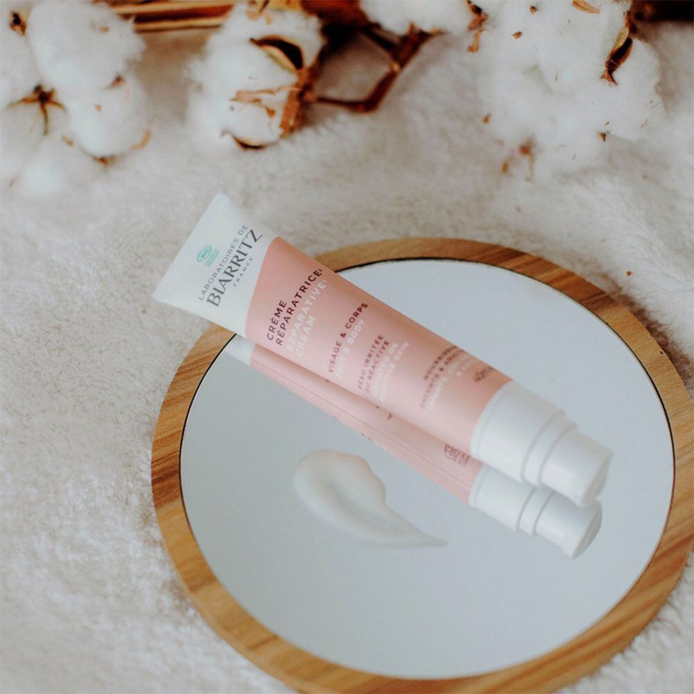 Creme-Tube auf der Schulter. Rosa-weißes Design. Text: Crème Réparatrice, Visage & Corps. Aufschrift: Peau irritée, abîmée, réactive.