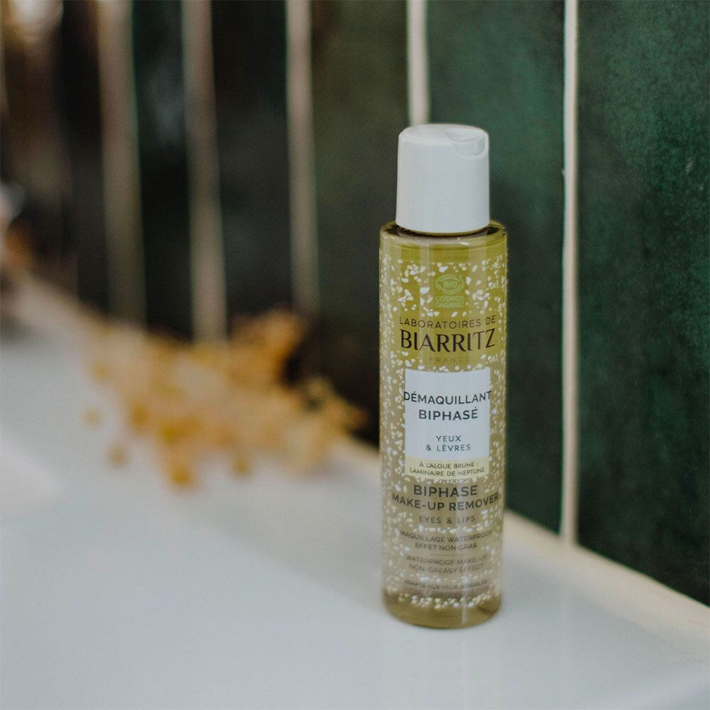 Eine Flasche Démaquillant Biphasé Yeux & Lèvres Bio steht auf einer weißen Oberfläche. Grüner Hintergrund.