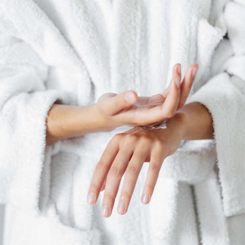 Person trägt Creme auf Hände auf. Weißer Bademantel. Nahaufnahme der Hände.