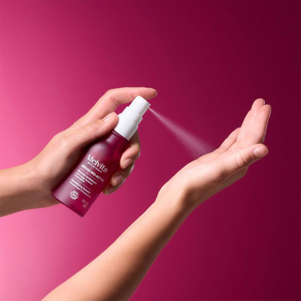 Hände sprühen Flüssigkeit aus einer roten Flasche. Aufschrift: Melvita Argan Bio-Active.