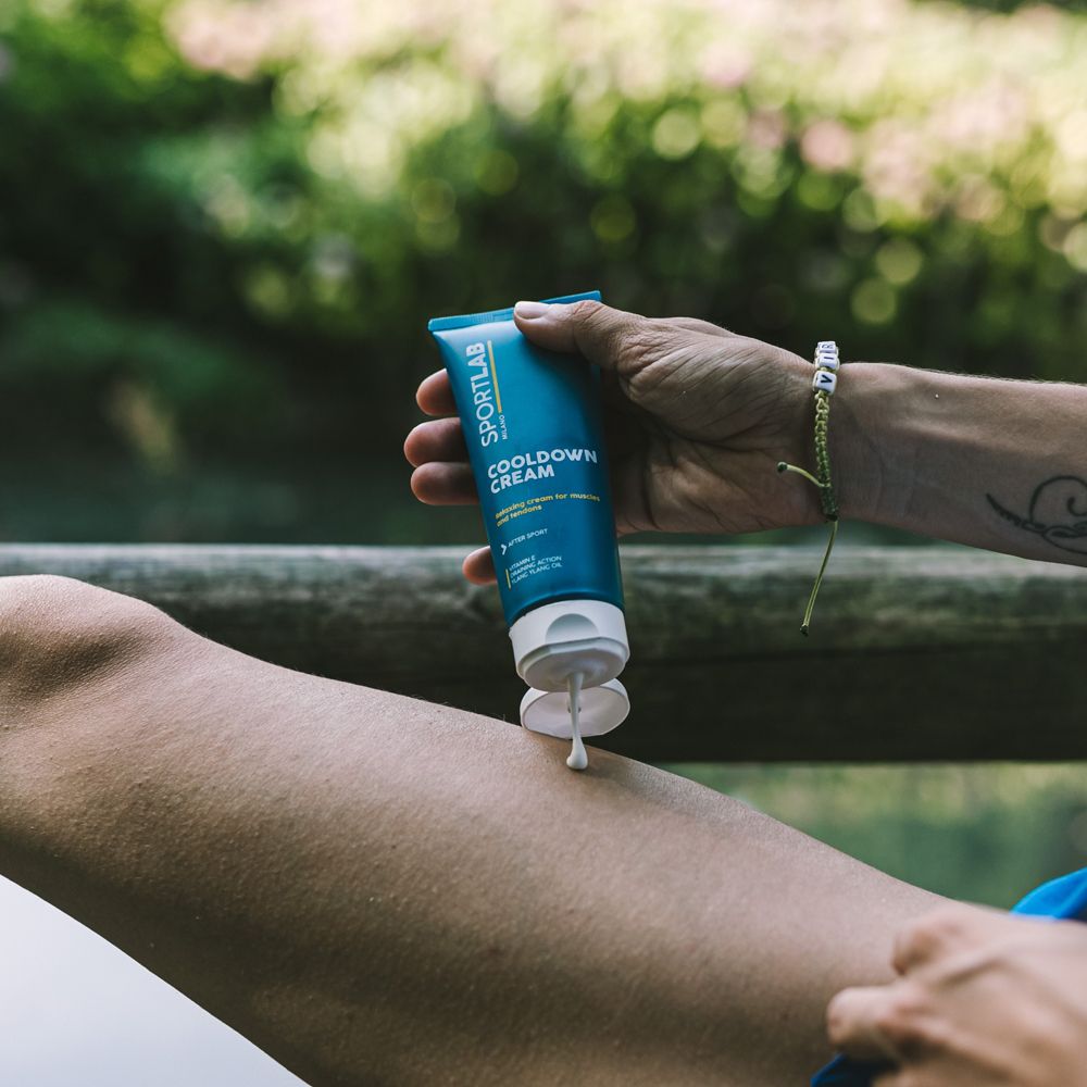 Persona applica crema sulla gamba. Testo: SPORTLAB Milano, COOLDOWN CREAM. Abbigliamento sportivo. Ringhiera in legno sullo sfondo.