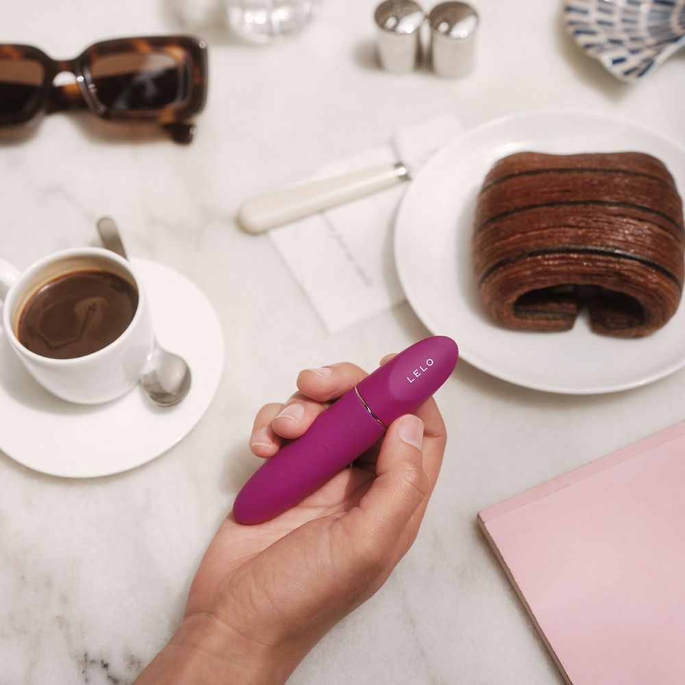 Person hält ein pinkfarbenes Wellness-Produkt. Kaffee, Croissant und Brille im Hintergrund.