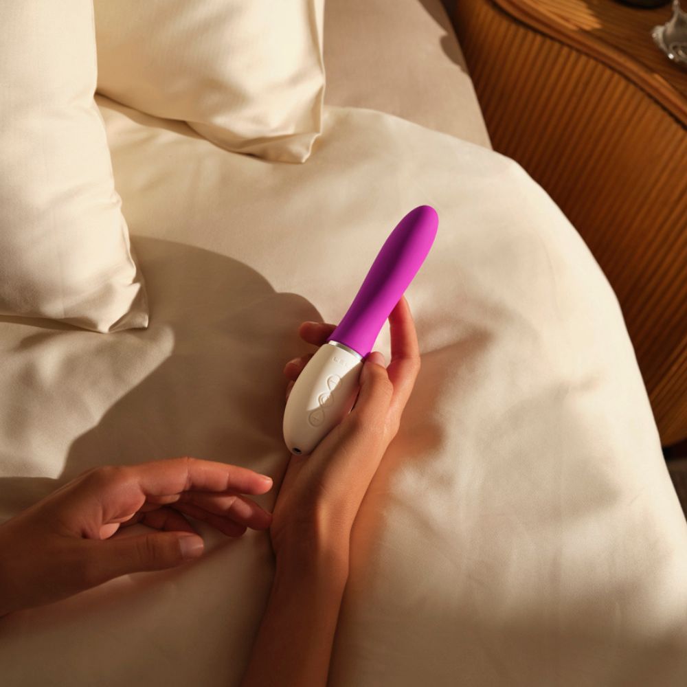 Person hält Vibrator. Weiß-lila. Auf Bettwäsche. LELO-Logo.