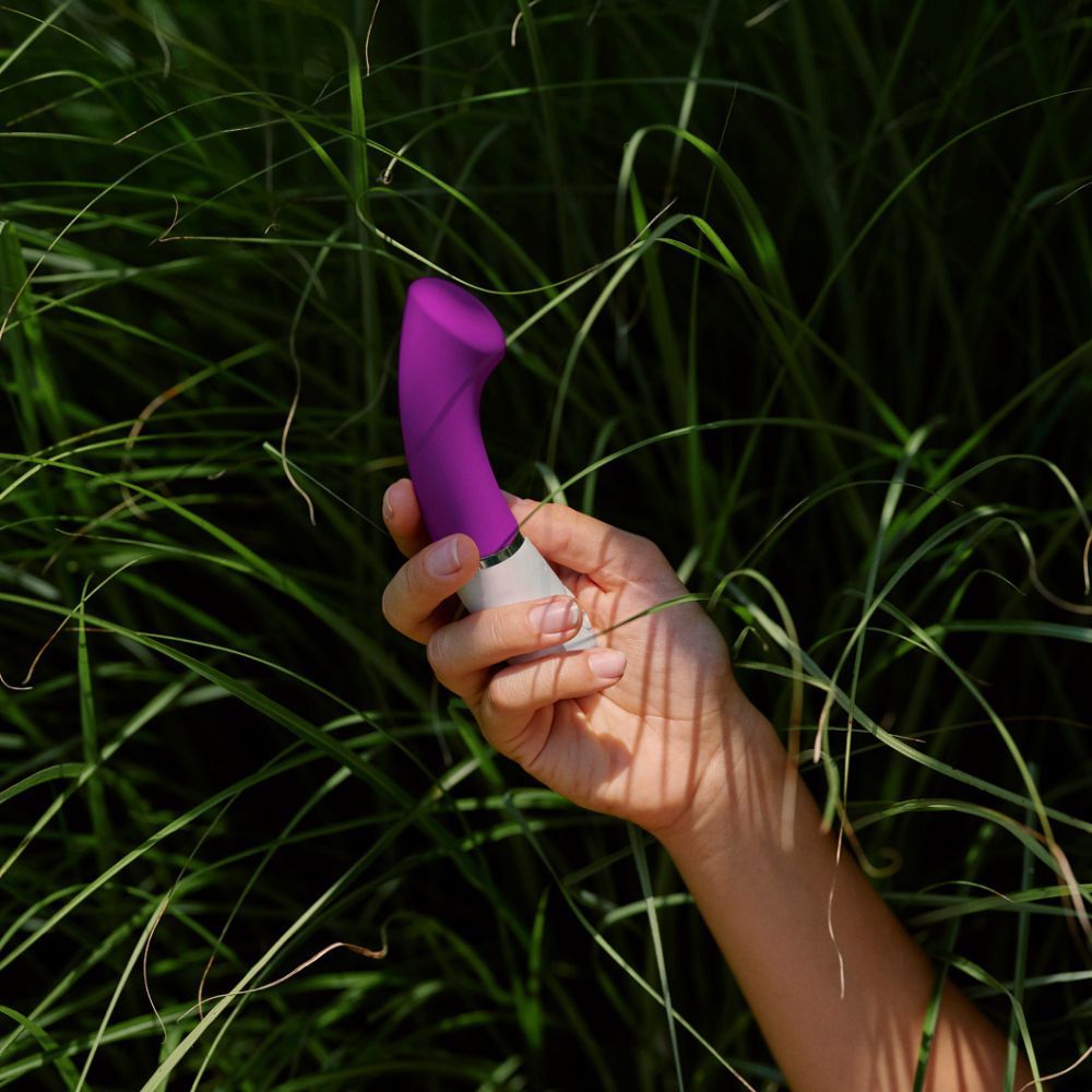 Hand hält lila-weißen Vibrator in hohem Gras. Gebogene Form.