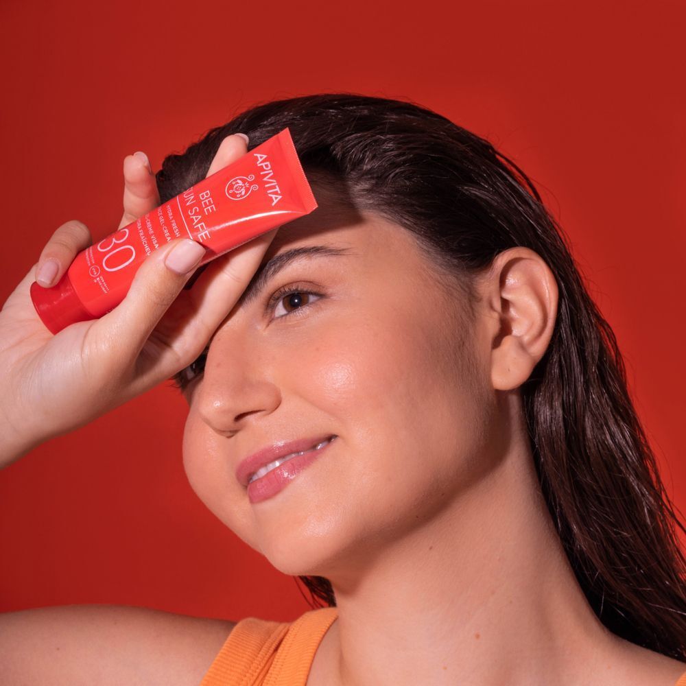 Una donna tiene un tubetto rosso APIVITA Bee Sun Safe Hydra Fresh Face Gel-Cream SPF 30 davanti alla fronte.