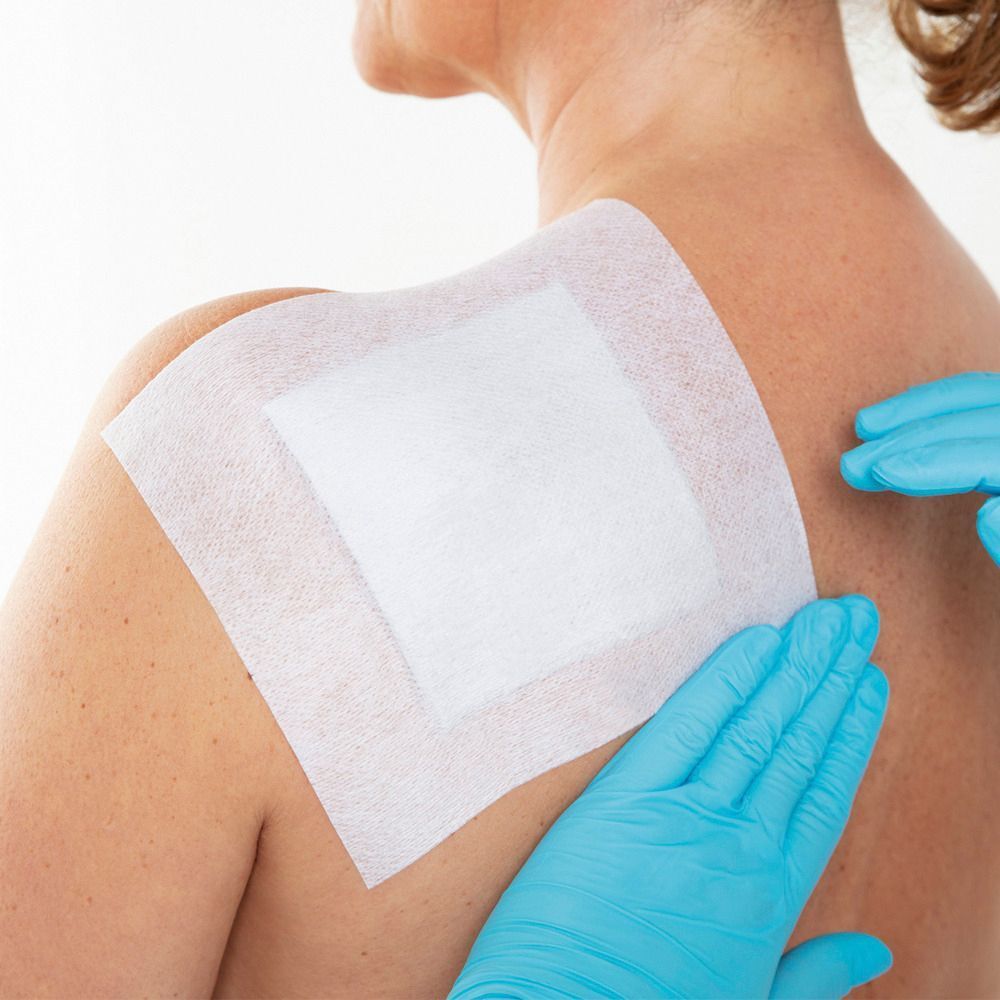 Leukoplast Fixomull Stretch applicato su una spalla. Mani con guanti blu che tengono la medicazione.