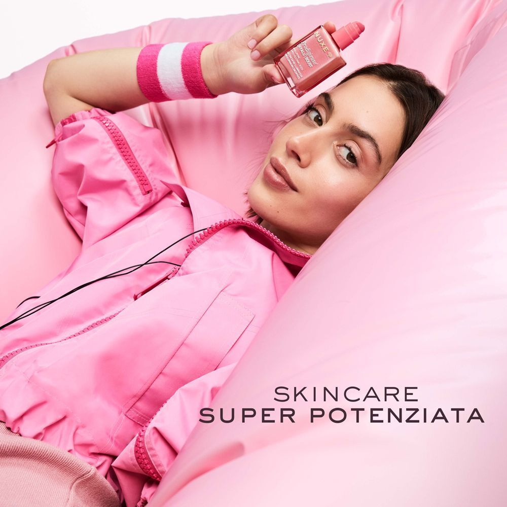 Donna sdraiata su cuscini rosa, tiene in mano un flacone Nuxe Prodigieuse [Hyalu] Boost.