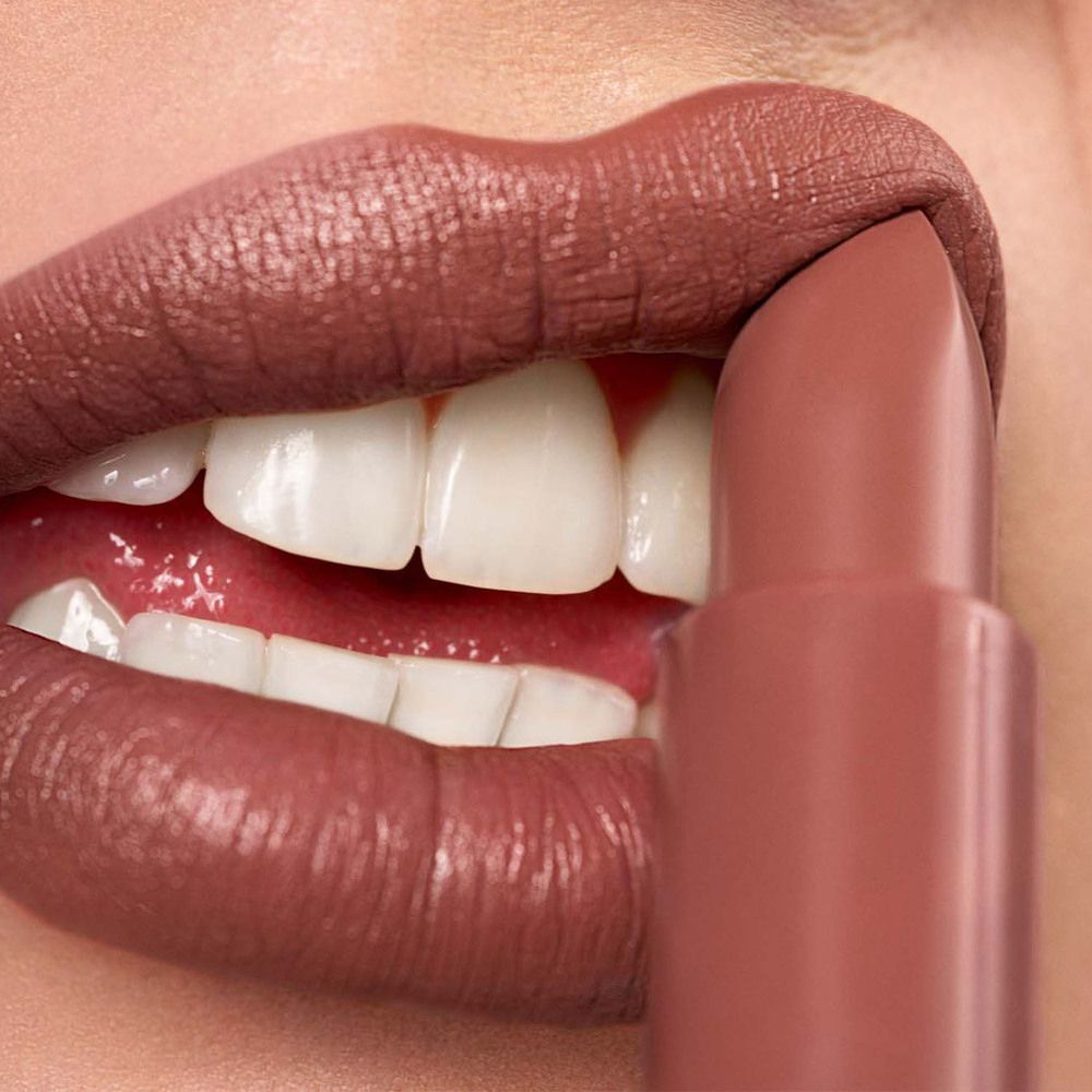 Un rossetto opaco color tortora viene applicato sulle labbra, i denti sono visibili.