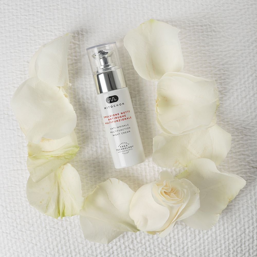 Flacone di crema notte multifunzionale antirughe Mitochon, circondato da petali di rosa bianchi.