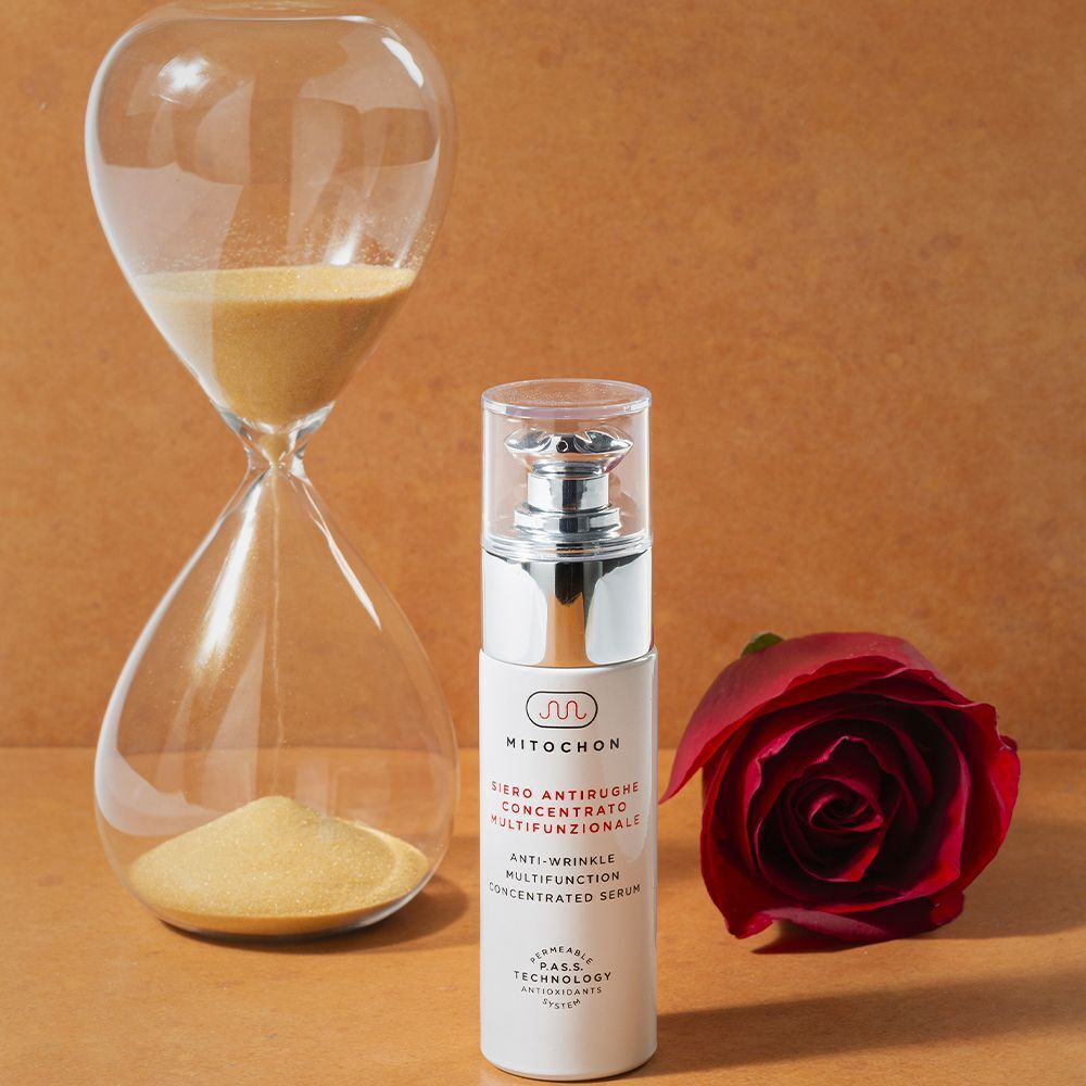 Flacone dispenser accanto a clessidra e rosa rossa, Mitochon Siero Concentrato Anti-Rughe Multifunzionale.