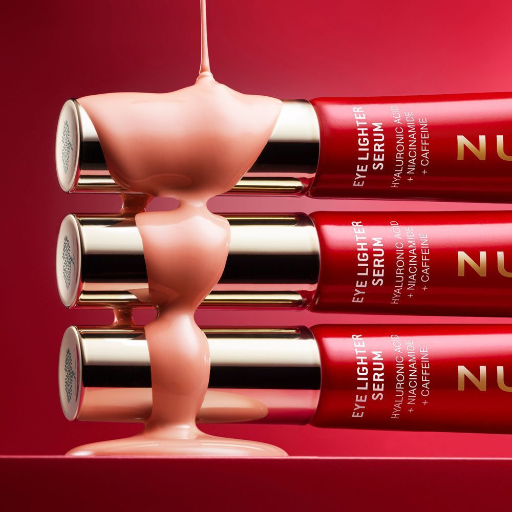 Siero che cola su tre tubetti rossi impilati Nuxe Eye Lighter Serum.