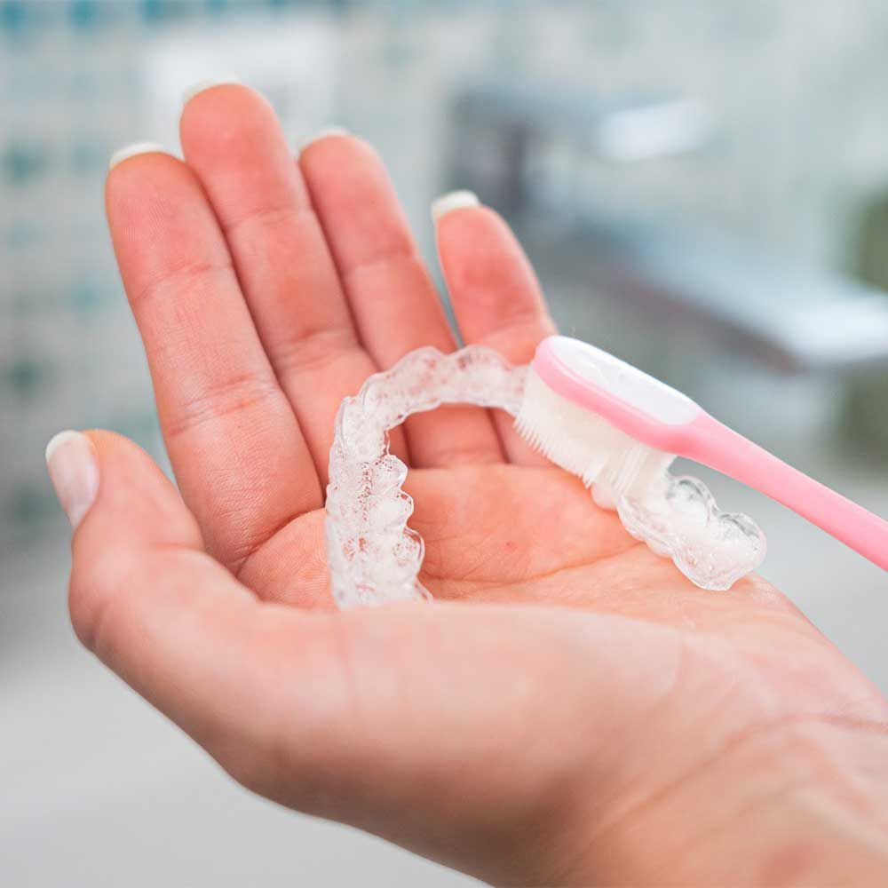 Una mano tiene un apparecchio trasparente per denti, mentre uno spazzolino rosa lo pulisce.