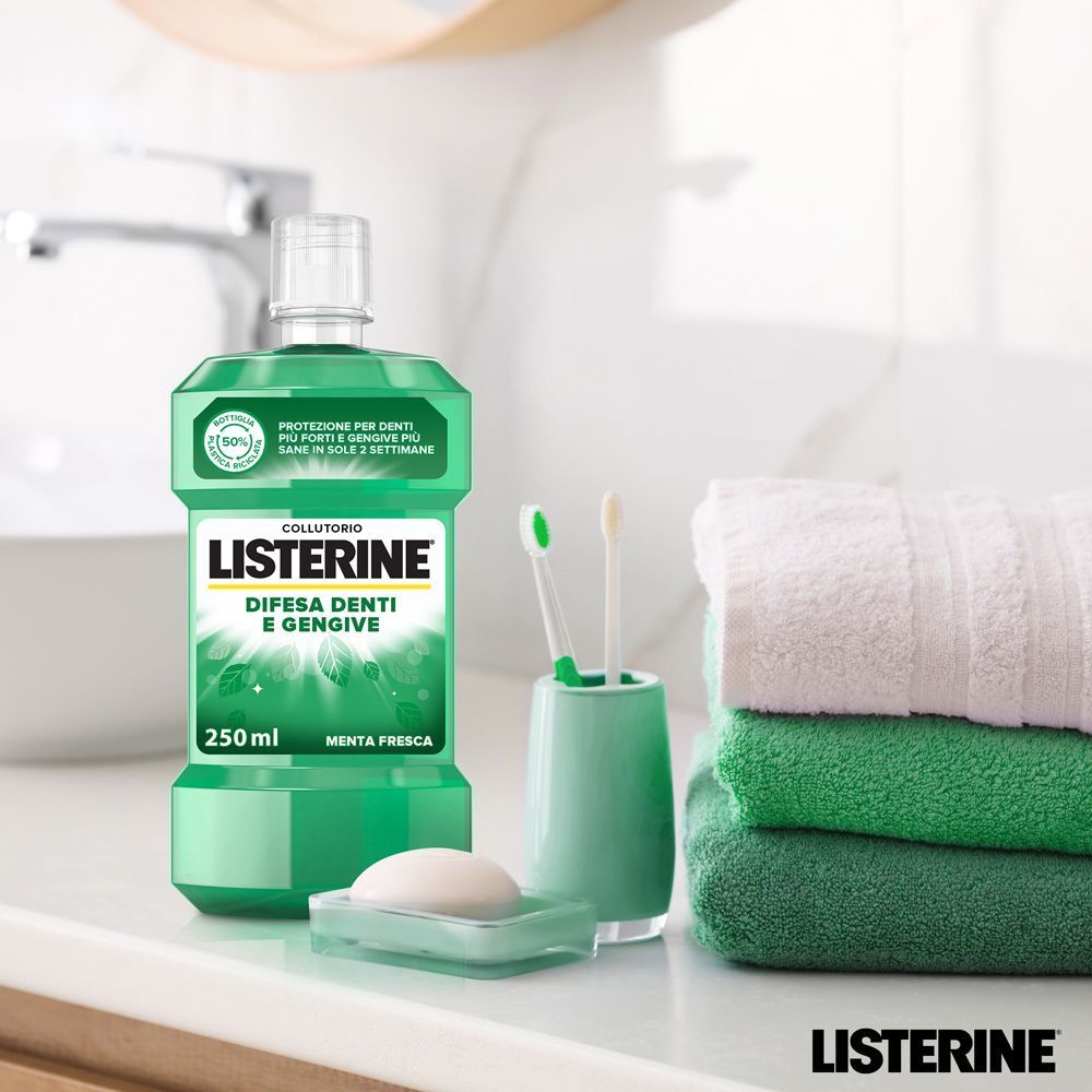 Flacone di collutorio Listerine verde da 250ml, etichetta "Difesa Denti e Gengive". Accanto, portasapone, portaspazzolino con due spazzolini e asciugamani impilati.