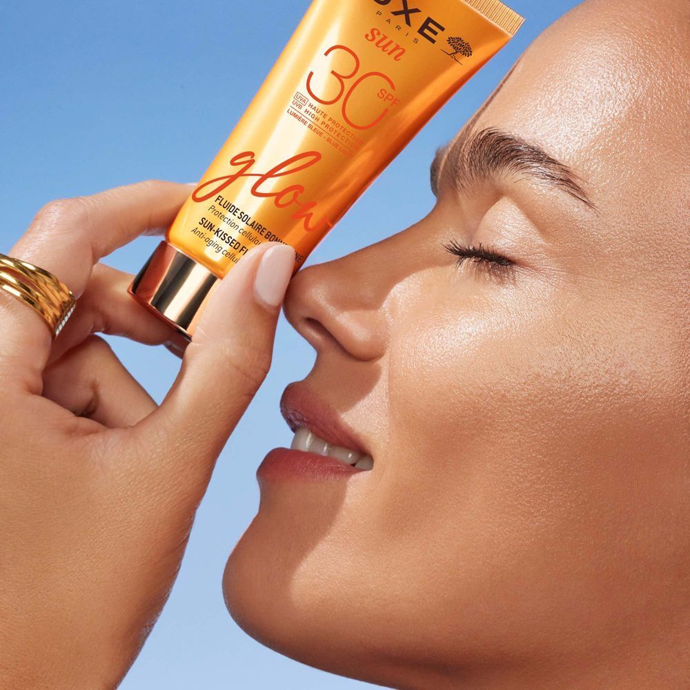 Una donna tiene un tubetto di Nuxe Sun Fluido Solare Viso Glow SPF 30 vicino al viso.