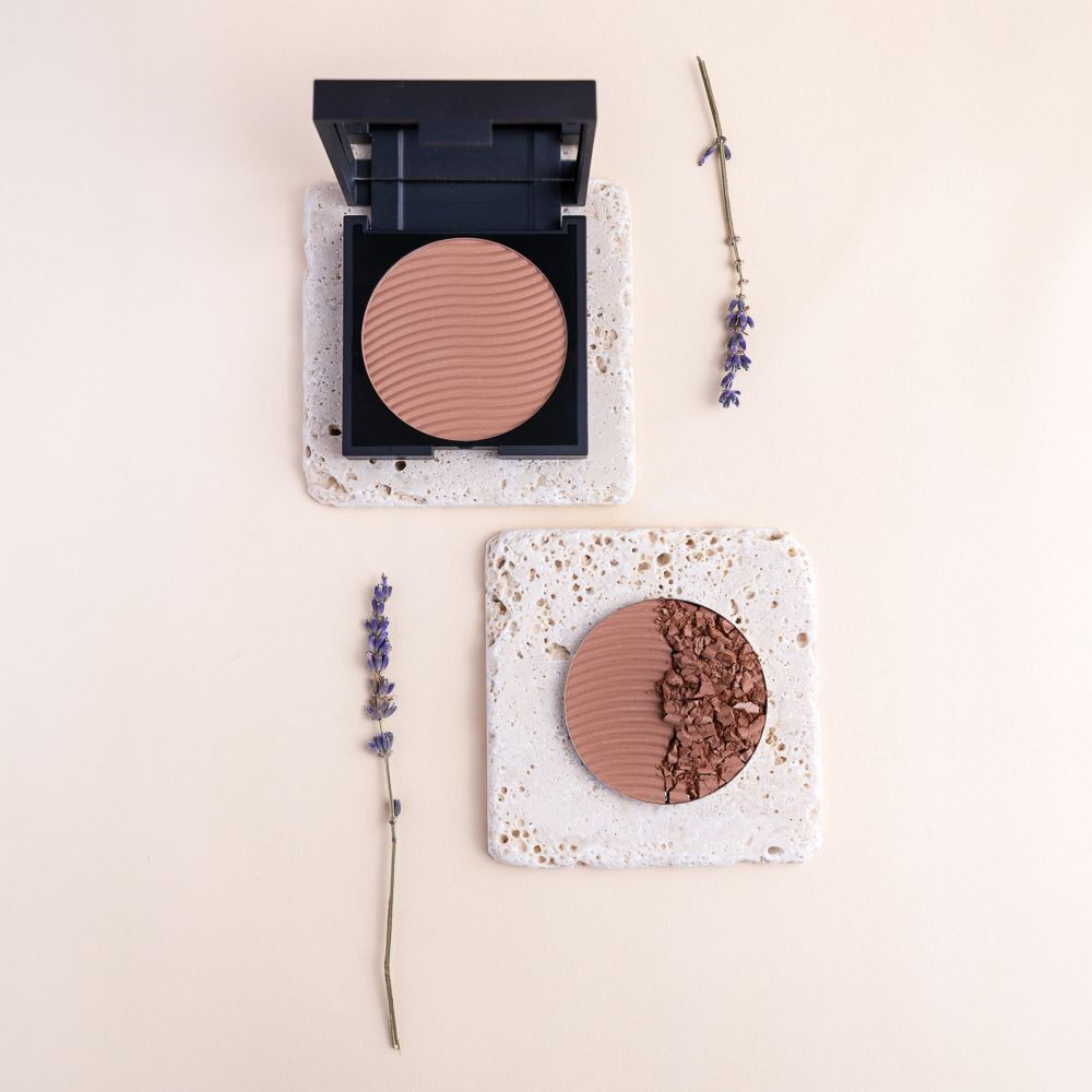Un bronzer compatto quadrato aperto, con cipria pressata rotonda color nocciola chiaro e motivo a onde.