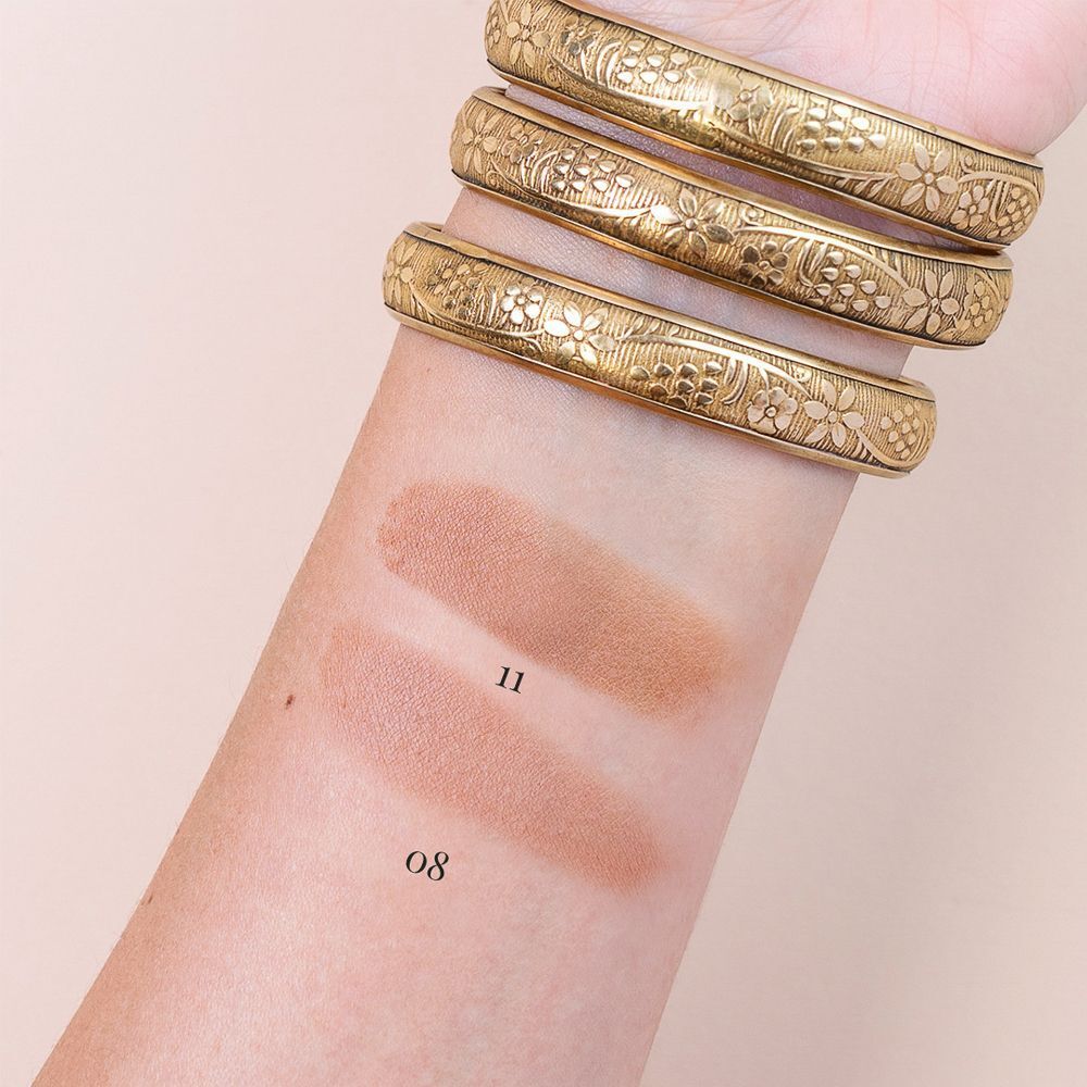 Due campioni di trucco marrone su un avambraccio, numerati 08 e 11. Bracciali dorati visibili.