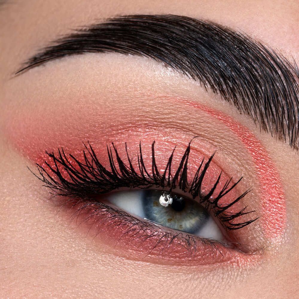 Un occhio con ombretto corallo, eyeliner e mascara nero volumizzante. Iride blu visibile.
