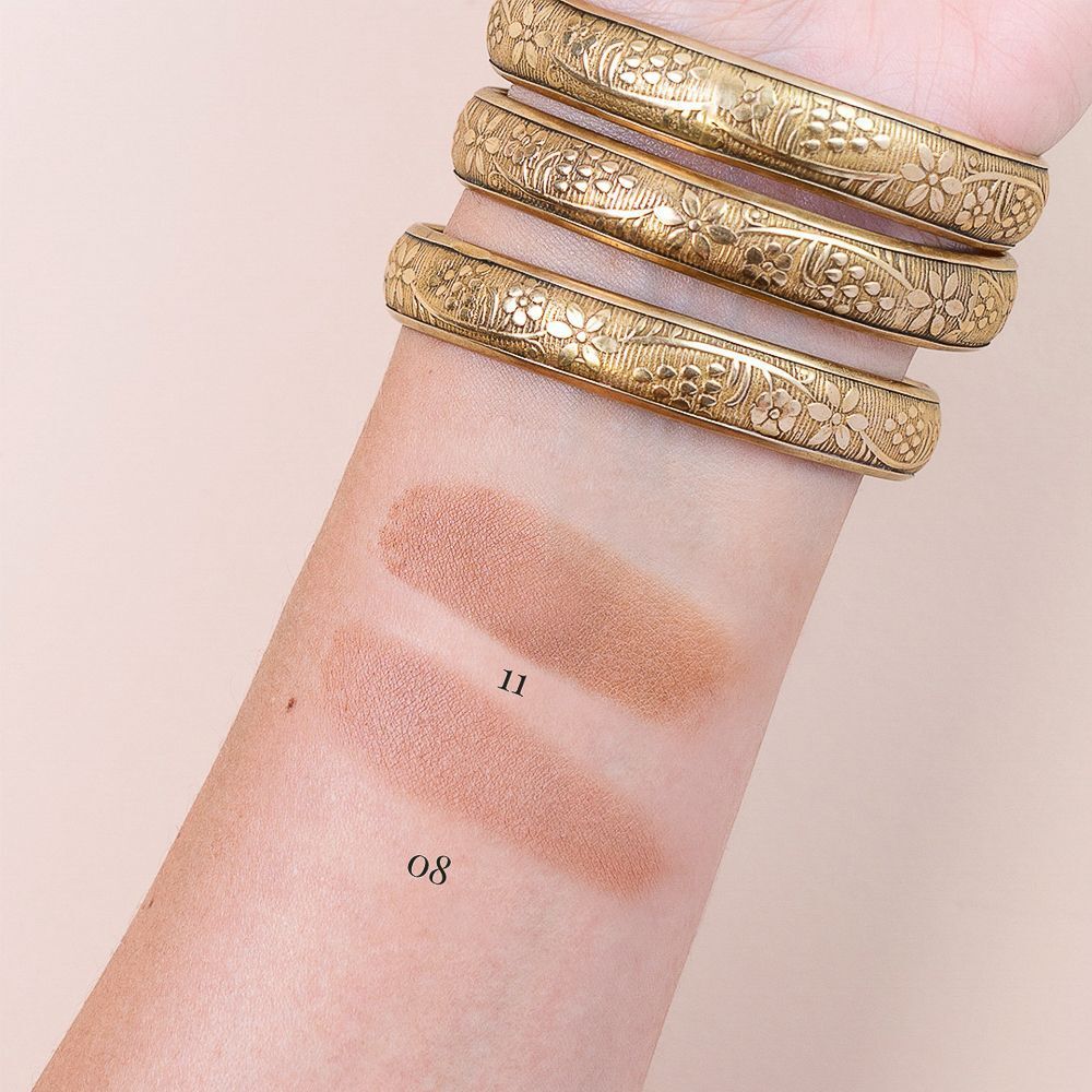 Due campioni di trucco marroni su un avambraccio, numerati 08 e 11. Bracciali dorati visibili.