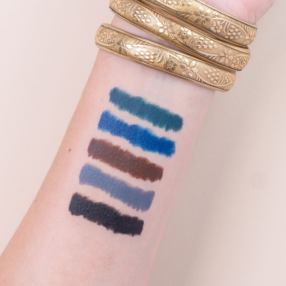 Cinque campioni di eyeliner su un avambraccio: verde scuro, blu reale, marrone, grigio, nero. Bracciali dorati al polso.