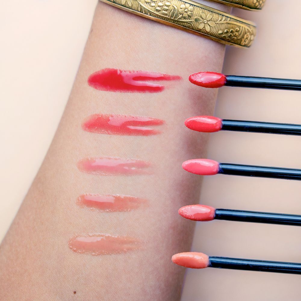 Cinque campioni di prodotto labbra brillante su un avambraccio, nei toni del rosso, rosa e nude. Applicatori con prodotto accanto.