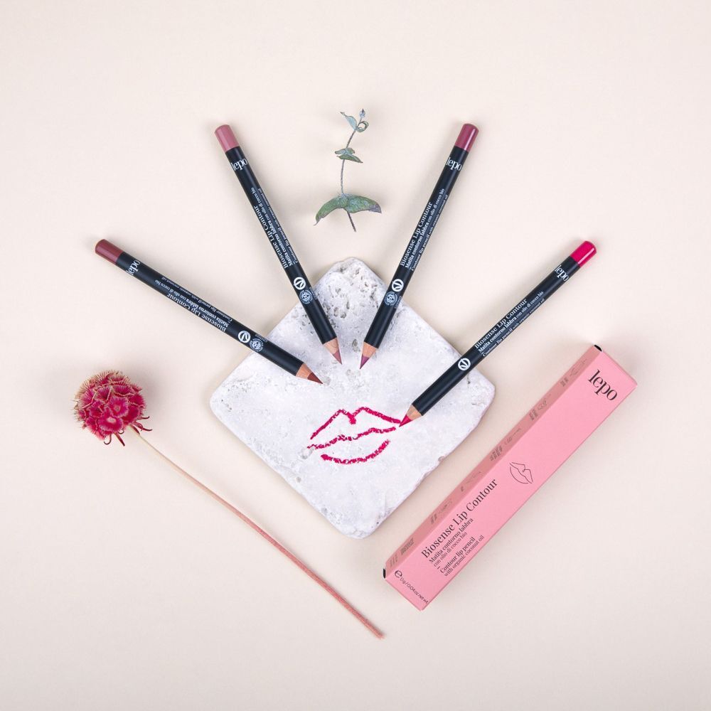 Quattro matite labbra in varie tonalità di rosa sono disposte attorno a una pietra bianca con labbra disegnate. Una confezione rosa è posizionata accanto.