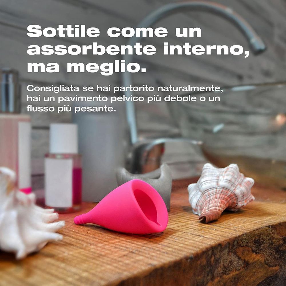 Coppetta mestruale rosa su mensola in legno accanto a conchiglie.