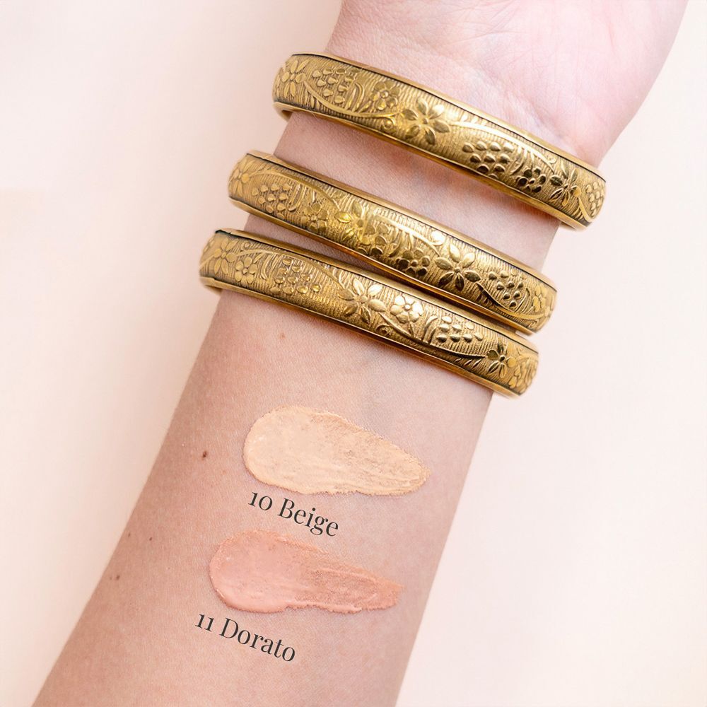 Due campioni di fondotinta su un braccio: 10 Beige e 11 Dorato. Bracciali dorati con motivo floreale.