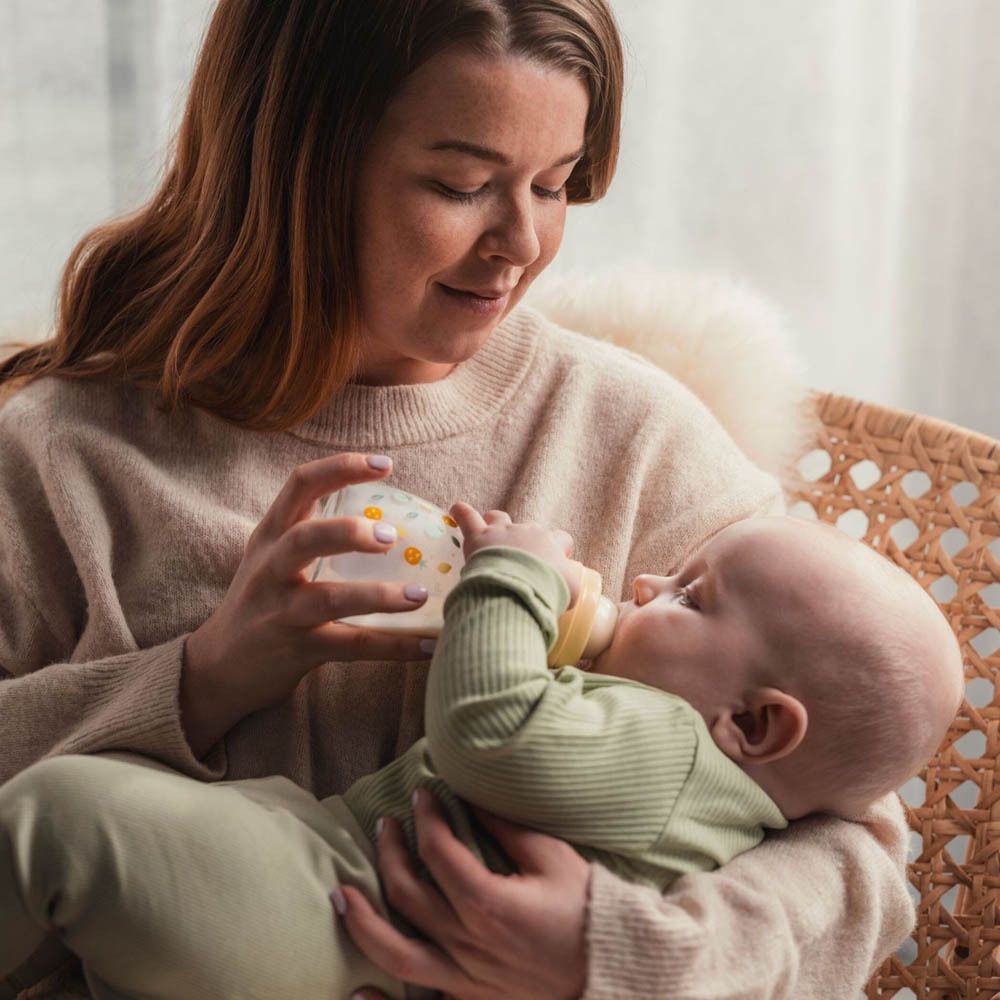 Eine Frau füttert ein Baby mit einer Flasche. Die Flasche hat ein Muster aus kleinen gelben Blumen.